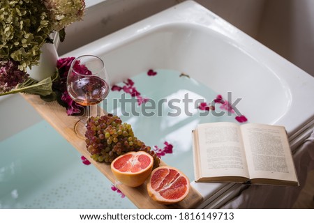 Bath tub with flower petals, grapefruit slices, bunch of grapes, a glass of wine, opened book and hydrangea bouquet. Organic spa relaxation preparation