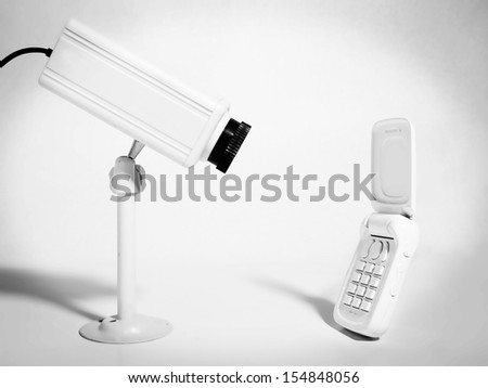 A security camera stares at a white cell phone against a white background.