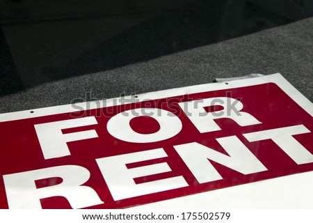 A For Rent Sign lies in a window of an empty window.