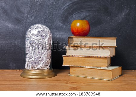 A glass dome full of shredded paper sits on a wooden table with a stack of books and an apple.