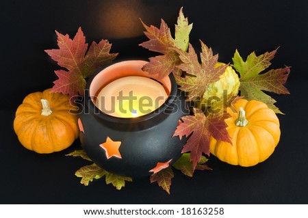 A candle in a cauldron with maple leaves and pumpkins.  The reflected light  from the flame casts an orange spooky glow on the  background.