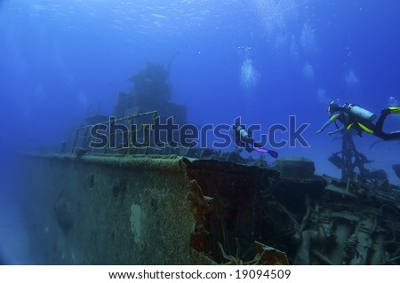 Two divers are swimming towards the superstructure of a shipwreck. Tangled wreckage in the foreground. Space for copy above.