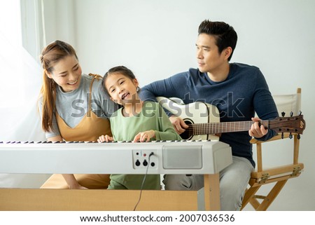 Happy little Asian family daughter playing piano with mother and father play guitar at home, Mother teaching daughter to play piano,They play and sing songs. concept for family relationship.