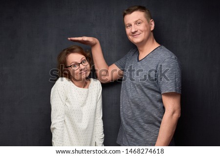 Studio waist-up shot of tall man smiling and showing with hand at height of short girl standing beside him and looking with perplexity at camera, over gray background. Variety of person's heights