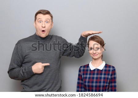 Studio shot of surprised tall man showing height of short woman, pointing at her with finger, both are standing over gray background. Concept of diversity of people's heights, tall and short persons