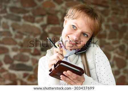 Young girl is smiling, talking by phone and writing down some information in pencil on paper, at stone wall background