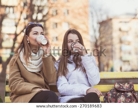 Mother and daughter sitting in autumn or spring in the park on a bench outdoors, drink coffee or tea or relax breakfast snack.