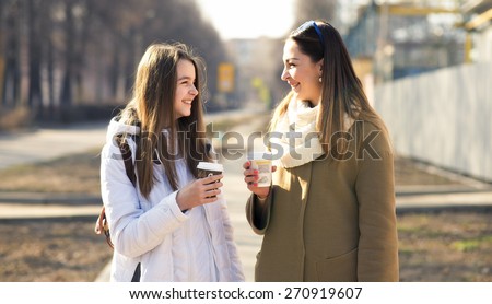 Mother and daughter talking, laughing and smiling on the street, drinking coffee in cups, in the fall or spring day, happy family.