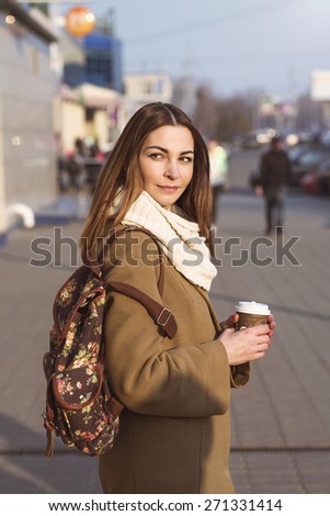 Portrait of a beautiful young brunette woman looking into the distance, in a coat and a scarf, standing in the street in spring or autumn. In his hand a cup of hot tea or coffee, fashion shoulder bag.
