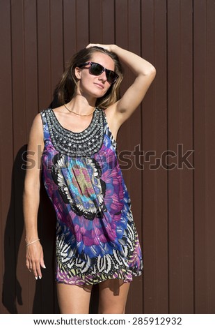 Beautiful young woman with glasses standing in front of a brown fence, fashion style bright summer dress.