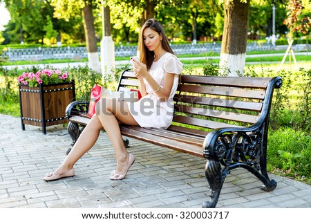 Beautiful girl sitting on a bench, brunette in a pink dress, fashion life style with your phone writes a message on social networks, sunny day in the park, relaxing, after work business woman.