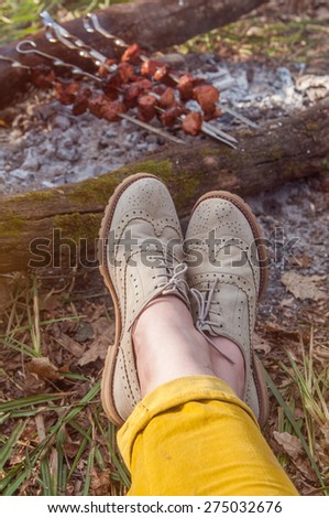 Recreation scene: woman feet in light brogues by camp fire in spring forest, grilled meat in the background