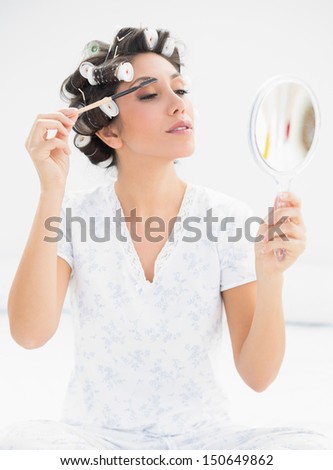 Happy brunette in hair rollers looking in hand mirror and brushing her eyebrows at home in bedroom