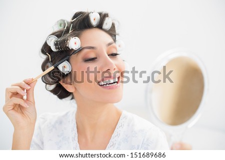 Smiling brunette in hair rollers looking in hand mirror and brushing her eyebrows at home in bedroom
