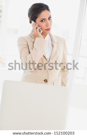 Frowning businesswoman standing behind her chair on the phone looking at camera in her office