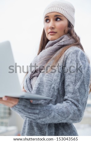 Thoughtful woman with winter clothes on using her laptop outdoors on a cold grey day