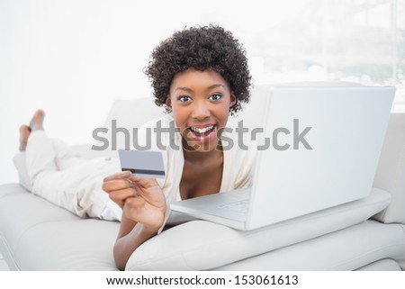 Smiling pretty brunette shopping online using her laptop lying on cosy sofa