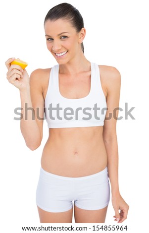 Smiling woman in sportswear holding slice of orange looking at camera against white background