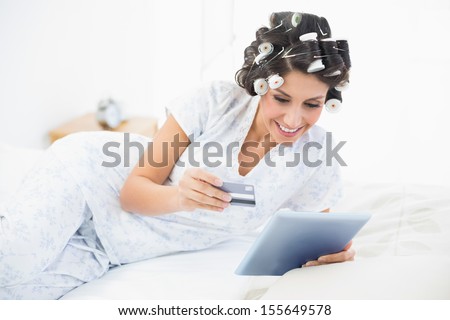 Excited brunette in hair rollers lying on her bed using her tablet to shop online in bedroom at home