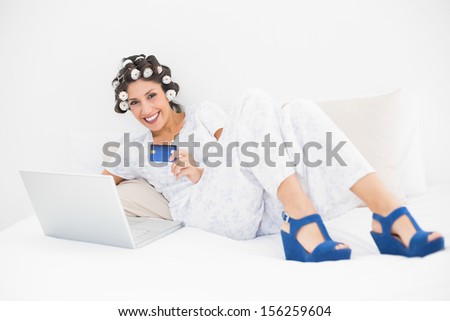 Brunette in hair rollers and wedge shoes using her laptop to shop online on bed in bedroom at home