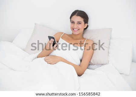 Relaxed young brown haired model in white pajamas using a mobile phone in bright bedroom
