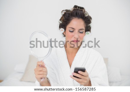 Smiling natural brunette holding mirror and smartphone in bedroom