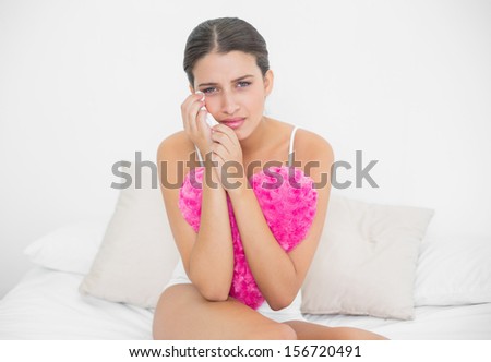 Sad young brown haired model in white pajamas wiping her tears in bright bedroom