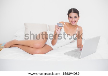 Relaxed young brown haired model in white pajamas shopping online with her laptop in bright bedroom