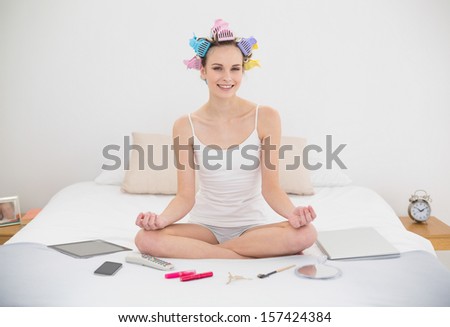 Content natural brown haired woman in hair curlers relaxing in lotus position in bright bedroom