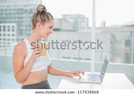 Cheerful sporty blonde using laptop holding coffee in bright room