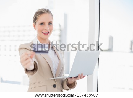 Cheerful blonde businesswoman in bright office showing credit card holding laptop