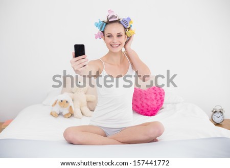 Smiling natural brown haired woman in hair curlers taking a picture of herself with mobile phone in bright bedroom