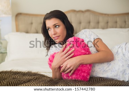 Calm pretty brown haired woman hugging a heart shaped pillow in a chic bedroom