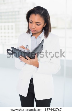 Thoughtful young dark haired businesswoman taking notes on her schedule in bright office