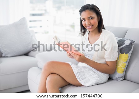 Amused young dark haired woman in white clothes reading a book in a living room
