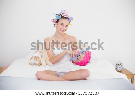 Amused natural brown haired woman in hair curlers using a tablet pc in bright bedroom