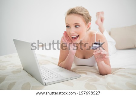 Natural excited blonde holding credit card and looking at laptop in bright bedroom