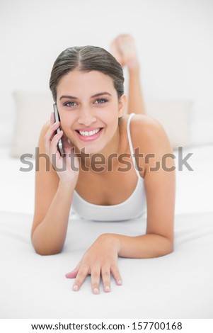 Casual young brown haired model in white pajamas making a phone call in bright bedroom