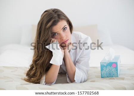 Sad casual brown haired woman in white pajamas crying on her bed in a bright bedroom