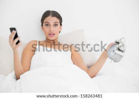Astonished young brown haired model in white pajamas holding a mobile phone and an alarm clock in bright bedroom