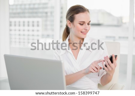 Happy businesswoman using laptop and texting on smartphone in her office