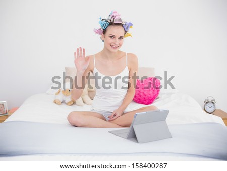 Joyful natural brown haired woman in hair curlers chatting online with a tablet pc in bright bedroom