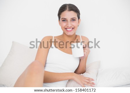 Cheerful young brown haired model in white pajamas enjoying coffee in bright bedroom