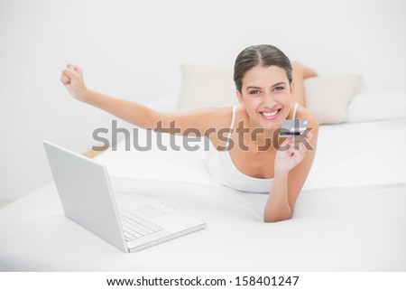Excited young brown haired model in white pajamas shopping online with her laptop in bright bedroom