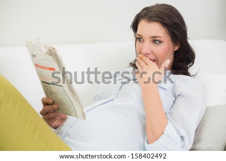 Surprised pregnant brown haired woman reading a newspaper in a bright living room
