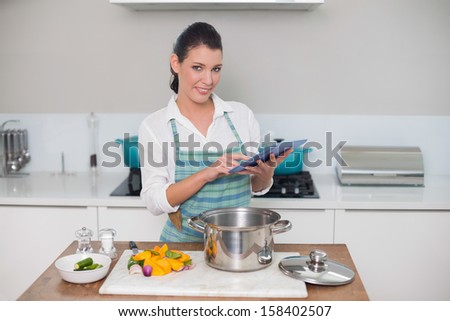 Cheerful gorgeous woman wearing apron using tablet while cooking in bright kitchen