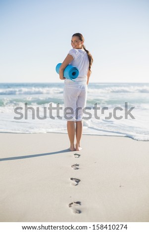 Rear view of gorgeous woman on the beach holding exercise mat looking over shoulder at camera
