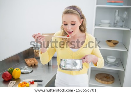 Content cute blonde tasting food from wooden spoon in bright kitchen