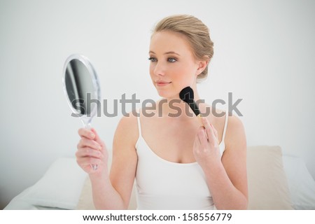 Natural content blonde holding mirror and using brush in bright bedroom