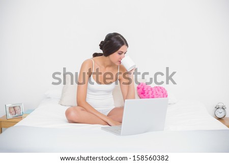 Peaceful young brown haired model in white pajamas drinking coffee in bright bedroom